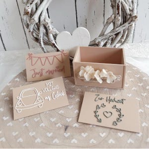 Könnte beinhalten: Eine hellbeige Holzbox mit weißen Blumenakzenten, begleitet von drei rechteckigen Schildern. Ein Schild zeigt ein Wimpeldesign und den Text "Just married", ein weiteres zeigt den Umriss eines Autos mit "MIT VOLLGAS ins Glück" und das dritte zeigt "Zur Hochzeit" mit einem Herz und einem Blattkranz.