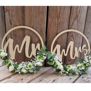 May include: Two wooden wedding signs, one reading "Mr" and the other "Mrs", each encircled with a wooden ring. The signs are decorated with white roses, green foliage, and white accents, set against a wooden background.