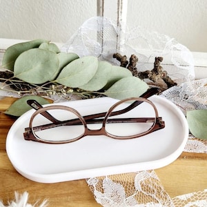 May include: A white oval ceramic tray with a pair of brown eyeglasses resting on it. The tray is on a wooden surface with white lace fabric.