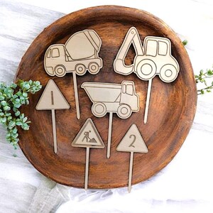 May include: Wooden construction-themed cake toppers. The set includes a cement mixer, excavator, dump truck, and road signs with numbers 1 and 2, and a construction worker symbol. The toppers are arranged on a wooden plate.