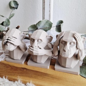 May include: Three beige monkey sculptures, each in a different pose: covering eyes, mouth, and ears. The geometric design gives a modern look. They sit on square bases, arranged on a wooden surface, with dried greenery in the background. A decorative home accent.