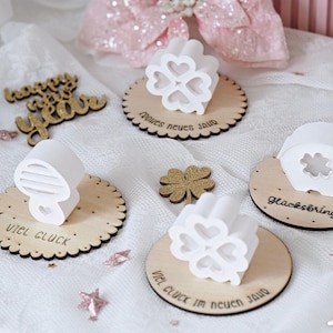 May include: White clover-shaped decorations and wooden coasters with text are arranged on a white surface. A gold glitter clover and a "Happy New Year" sign are also present. The coasters have scalloped edges.