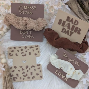 Peut inclure: Quatre chouchous de cheveux bruns et beiges avec des motifs différents. Les chouchous sont présentés sur des cartes brunes et beiges avec du texte qui dit "Messy Hair Don't Care", "Bad Hair Day", "You Got The Look" et un motif léopard.