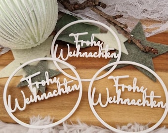 Puede incluir: Tres adornos circulares blancos con la frase alemana "Frohe Weihnachten". Se colocan sobre una superficie de madera con decoraciones en forma de estrella verde y una rama decorativa. Una gran esfera blanca texturizada está en el fondo.