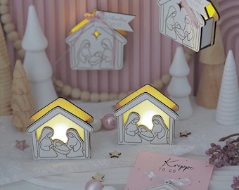 May include: Wooden nativity scene ornaments in a house shape, depicting Mary, Joseph, and baby Jesus. The roofs are yellow and the interiors are illuminated. Some ornaments are suspended with ribbons. The scene is set against a pink backdrop.