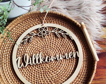 Puede incluir: Un letrero redondo de madera con la palabra "Willkommen" en escritura cursiva. El letrero está decorado con un diseño floral y de hojas y está colgado sobre una cesta tejida. Una clavija de madera descansa sobre la cesta.
