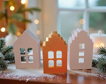 May include: Three decorative house-shaped candle holders in a row. The houses are painted in shades of white and orange. Each house has square cut-out windows. The houses are sitting on a wooden surface with a dusting of white powder.