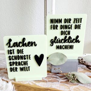 May include: Two light green signs with black lettering. The first sign says "Lachen ist die schönste Sprache der Welt" with a black heart. The second sign says "Nimm dir Zeit für Dinge die dich glücklich machen!"