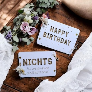 May include: Two white wooden signs with gold lettering. The top sign says "Happy Birthday" and the bottom sign says "Nichts So, wie du es dir gew&uuml;nscht hast".