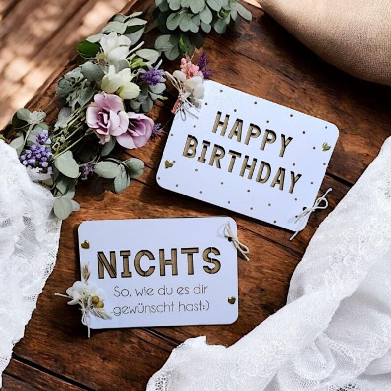 May include: Two white wooden signs with gold lettering. The top sign says "Happy Birthday" and the bottom sign says "Nichts So, wie du es dir gew&uuml;nscht hast".