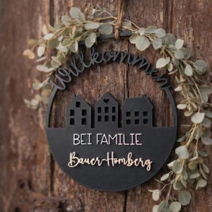 May include: A black circular welcome sign with the word "Willkommen" cut out at the top. Below are three house silhouettes and the words "BEI FAMILIE Bauer-Homberg" in white and gold. The sign is decorated with a green leafy garland.