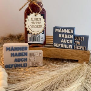 May include: A dark liquid bottle with a scalloped wooden tag reading KLEINES MÄNNER GESCHENK, accompanied by two light-colored blocks with blue text reading MÄNNER HABEN AUCH GEFÜHLE! HUNGER ZUM BEISPIEL and two blue blocks with white text reading MÄNNER HABEN AUCH GEFÜHLE! DURST ZUM BEISPIEL.