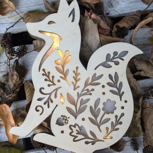 May include: A wooden fox-shaped decorative light with floral cutouts. The fox is made of light-coloured wood, with warm light emanating from within the cut-out designs. The design includes leaves, flowers, and an acorn. The fox is placed on a bed of dried leaves.
