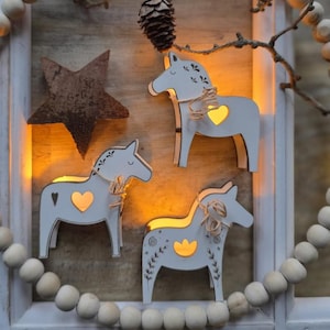 May include: Light grey wooden Dala horse ornaments with heart-shaped cutouts that glow. Each horse has floral accents and twine detailing. A rustic star and wooden beads frame the scene. The horses are approximately 15 cm in height.