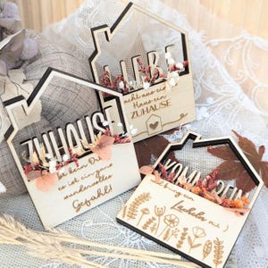 May include: Three wooden house-shaped decorations with black outlines and floral accents. Each piece features a different German phrase, including "ZUHAUSE" and "LIEBE", and decorative floral arrangements.
