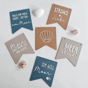 Könnte beinhalten: Eine Sammlung dekorativer Wimpel in Blau-, Braun- und Grautönen mit weißem Text und Muscheldesign. Die Wimpel zeigen Sätze wie "STRAND Liebe" und "ICH WILL Meer". Muscheln sind um die Wimpel herum verstreut.
