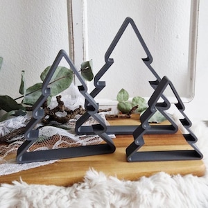 Pode incluir: Três decorações cinzentas de metal em forma de árvore de Natal de tamanhos diferentes. As árvores estão colocadas numa tábua de cortar de madeira. O fundo inclui vegetação e uma janela branca.