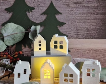 May include: A collection of small, white, house-shaped candle holders with cut-out windows. Some are lit from within, casting a warm glow. The background features green tree cutouts and a wooden texture.