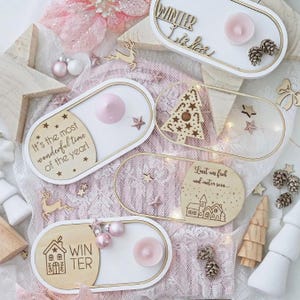 May include: A collection of decorative wooden trays with white and gold accents, each holding a small pink candle. Some trays feature text like "Winter Liebe" and "It's the most wonderful time of the year!" along with festive designs like stars and a Christmas tree.