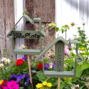 Könnte beinhalten: Zwei hausförmige Gartenstecker in Salbeigrün. Einer der Stecker trägt die Aufschrift "LIEBLINGS Platz", der andere "WILLKOMMEN". Die Stecker sind in einem Garten mit bunten Blumen platziert.