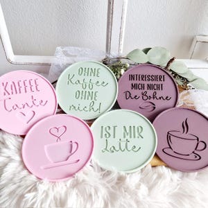 May include: Six round coasters with different designs and sayings. The coasters are made of a light pink, light green, and dark purple material. The sayings on the coasters are "Kaffee Tante", "Ohne Kaffee Ohne Mich!", "Interessiert Mich Nicht Die Bohne", "Ist Mir Latte", and two coasters with a coffee cup design.