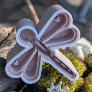 Może przedstawiać: Jasnoróżowa wykrawaczka do ciastek w kształcie ważki z białym obramowaniem. Wykrawaczka ma szczegółowy wzór z wycięciami w kształcie serca. Wykrawaczka jest wykonana z tworzywa sztucznego i leży na drewnianej powierzchni z zielonymi roślinami w tle.