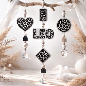 May include: A decorative mobile featuring shapes with a black and white leopard print design. The mobile includes a heart, rectangle, circle, and diamond shape, along with the word "LEO". Each shape is adorned with beads and a crystal.