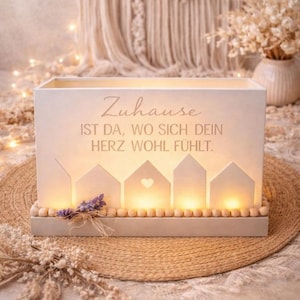 Könnte beinhalten: Rechteckige, cremefarbene Holz-Dekobox mit dem deutschen Text "Zuhause ist da, wo sich dein Herz wohl fühlt." Die Box hat hausförmige Aussparungen mit Lichtern und eine Holzperlenbasis. Ein kleines Bündel getrockneten Lavendels liegt auf der Basis.