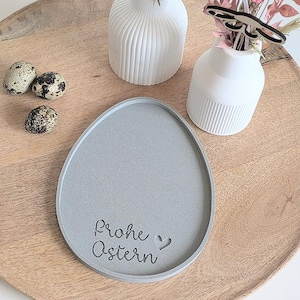 May include: A grey, egg-shaped decorative tray with the words "Frohe Ostern" and a heart symbol. The tray is on a wooden surface, with three speckled quail eggs and two white vases with flowers.