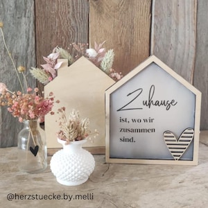May include: Wooden house-shaped decor with dried flowers and a small vase with a heart. A sign with the text "Zuhause ist, wo wir zusammen sind" and a heart is also present. The decor is set against a wooden background.