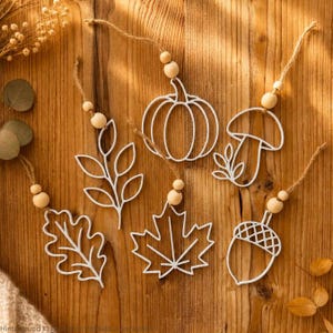 May include: Five white wire ornaments shaped like a pumpkin, a mushroom, an acorn, a maple leaf, and an oak leaf. Each ornament hangs from a jute twine string accented with two natural wooden beads, creating a set of seasonal hanging decorations.