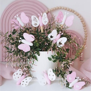 Puede incluir: Una maceta de cerámica blanca llena de vegetación y decoraciones de mariposas rosas y blancas. Las mariposas tienen varios patrones y están sobre palitos de madera. El fondo incluye un arco rosa y una guirnalda de cuentas de madera.