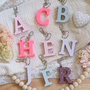 Può includere: Una collezione di portachiavi a forma di lettera in vari colori pastello, tra cui rosa, blu e viola. Ogni lettera è decorata con pois bianchi e attaccata a un moschettone argentato. Le lettere sono disposte su un tessuto color crema.