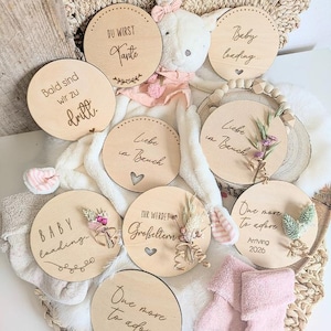 May include: A collection of round wooden plaques featuring engraved text including Baby loading, Du wirst Tante, Bald sind wir zu dritt, Liebe im Bauch, Ihr werdet Großeltern, and One more to adore Arriving 2026, decorated with dried flowers and small hearts.