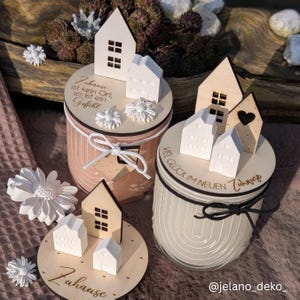 May include: Two white candle jars with wooden lids decorated with small wooden houses and the text "Zuhause ist kein Ort. es ist ein Gefuhl" and "VEL GLÜCK IM NEUEN Zuhause".
