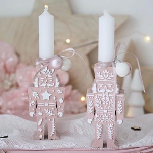 May include: Two pink and white nutcracker candle holders with white candles. The nutcrackers have detailed white designs and are decorated with pink and white ornaments. The background is soft pink and white.