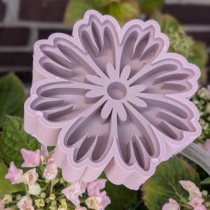 Puede incluir: Cortador de galletas en forma de flor de color lila claro, con múltiples capas y detalles de pétalos. El cortador está rodeado de hojas verdes y flores rosas, creando un tema natural y floral.