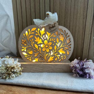 Op de afbeelding: Een houten pompoenvormige lamp met ingewikkelde bloemen- en bladeruitsparingen. Het warme licht schijnt door het ontwerp. Een klein label met de tekst "Herbst Moment" is bevestigd met een wit lint. Decoratieve bloemen staan op de voorgrond.