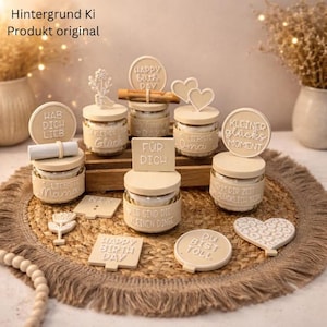 Puede incluir: Una colección de pequeños frascos de color beige con tapas de madera y adornos decorativos. Algunos adornos incluyen texto como "Happy Birthday" y "Liebste Mama". Los frascos están dispuestos sobre una estera tejida, lo que sugiere un regalo o decoración.
