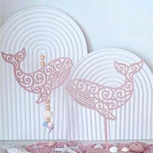May include: Two dusty pink wooden whale cutouts featuring intricate scrollwork patterns. One whale hangs from a string adorned with wooden beads, a small seashell, and a heart-shaped crystal, while the other is mounted on a vertical stand.