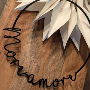 May include: A black metal wire wreath with the word "amore" in cursive script. The wreath is on a wooden surface, with a white paper starburst decoration in the background.
