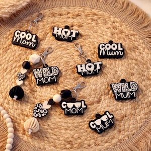 May include: A collection of black keychains with white text that reads "COOL Mom", "HOT Mum", and "WILD Mom". Some keychains have decorative beads in black and white. The keychains are displayed on a woven, circular mat.