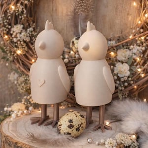 Puede incluir: Dos figuras decorativas de pájaros color crema con patas y picos marrones sobre una superficie de madera. Un pájaro tiene un detalle de corazón. Un huevo moteado descansa entre ellos. El fondo incluye una corona floral e iluminación suave.