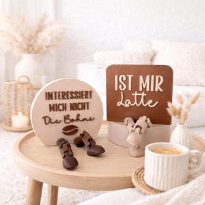 May include: A wooden coffee-themed display with the phrases "INTERESSIERT MICH NICHT Die Bohne" and "IST MIR Latte". The display includes a coffee cup, decorative wooden coffee beans, and a small vase with dried flowers. The items are arranged on a wooden table.
