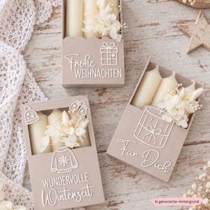 Op de afbeelding: Drie rechthoekige beige doosjes met crèmekleurige stompkaarsen en witte gedroogde bloemen. De doosjes zijn voorzien van witte lijntekeningen van een cadeau, een trui, een muts en wanten, met de Duitse tekst Frohe WEIHNACHTEN, WUNDERVOLLE Winterzeit en Für Dich.