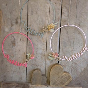 Op de afbeelding: Drie decoratieve kransen met het woord "Frühling" in script, elk met gedroogde bloemenaccenten. De kransen zijn roze, lichtgroen en lichtroze, hangend tegen een houten achtergrond. Twee houten hartvormige decoraties staan eronder.