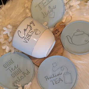 Op de afbeelding: Een lichtblauwe en witte mok met de woorden "Cozy Vibes" en een koffiekopje. De mok is omringd door lichtblauwe onderzetters met thee-gerelateerde ontwerpen en tekst, waaronder een theepot en de woorden "Positivei TEA".