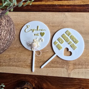 May include: Two white, round cake toppers with gold text. One says "Erzieherin mit" and has a small floral arrangement. The other says "Kleine Menschen brauchen so große Herzen" and has a heart cutout.