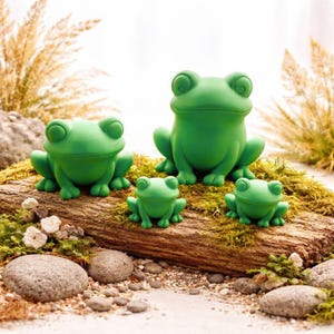 Könnte beinhalten: Vier grüne Froschfiguren in verschiedenen Größen sind auf einem mit Moos bedeckten Baumstamm angeordnet. Die Frösche haben große Augen und sitzen. Der Hintergrund umfasst hohes, trockenes Gras und kleine Steine.