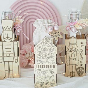 Könnte beinhalten: Weihnachtliche Geschenkboxen aus Holz mit Nussknacker-Design und Glasflaschen. Eine Box trägt die Aufschrift "LECKEREIEN" und eine andere "FROHE WEIHNACHTEN". Die Geschenke sind mit rosa Ornamenten und einem kleinen Beutel verziert.